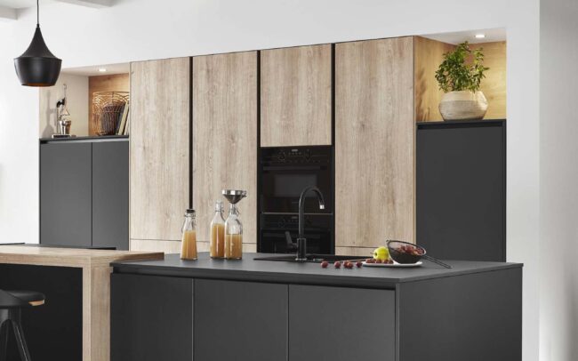 A modern Natura kitchen featuring Oak Montreal reproduction cabinets with a synchronized pore finish, paired with sleek matte black elements. The design includes a central island with integrated sink and minimalist decor, creating a cosy yet contemporary atmosphere. The realistic wood texture of the cabinets offers a natural look and feel, perfect for German kitchens at trade prices.