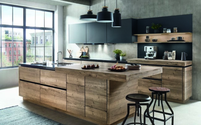 A chic urban kitchen featuring Structura's Havana oak reproduction finish, with spacious island and matching cabinetry highlighted by natural light from large windows. The modern aesthetic is complemented by dark countertops, sleek black shelving, and industrial-style stools, showcasing a blend of natural wood textures and contemporary design, ideal for German kitchens at trade prices.