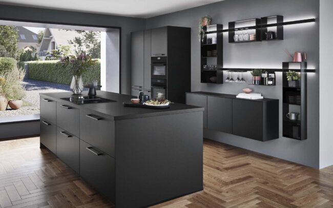 A sleek Touch kitchen in Black supermatt finish, featuring a large island, contemporary shelving, and integrated appliances. The space is styled with a combination of dark cabinets and light wood flooring, providing a modern, minimalist aesthetic. Large windows offer a view of the outdoors, enhancing the sophisticated, urban feel of the kitchen, ideal for German kitchens at trade prices.